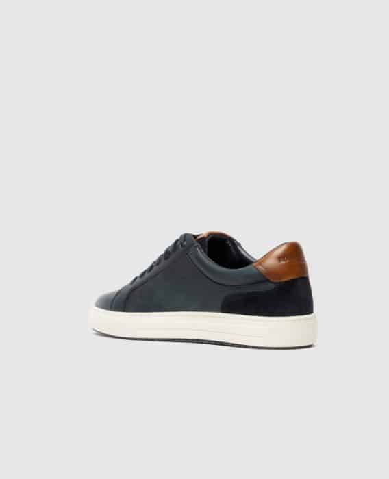 A black low-top sneaker with a white sole and brown leather accent on the heel, viewed from the back and side against a plain light gray background.