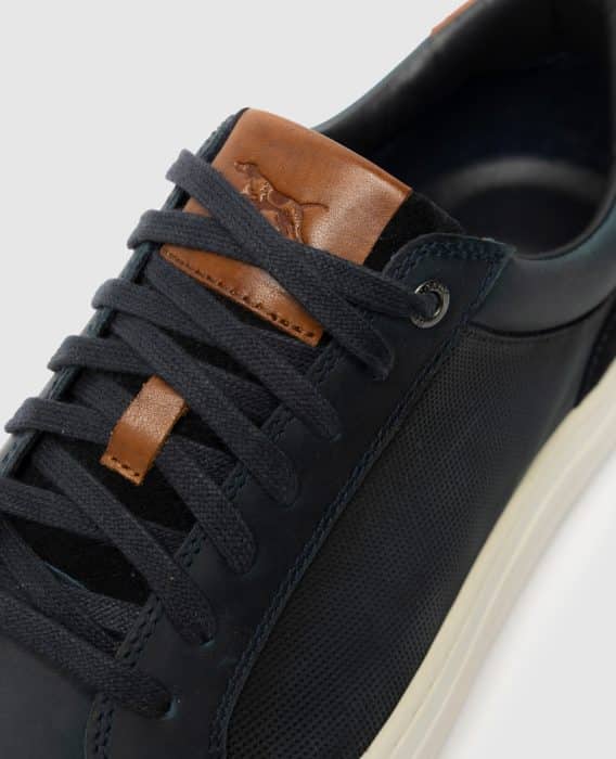 Close-up of a dark blue sneaker with black laces, brown leather accents on the tongue, and a subtle embossed animal logo. The shoe has a textured side panel and an off-white sole.