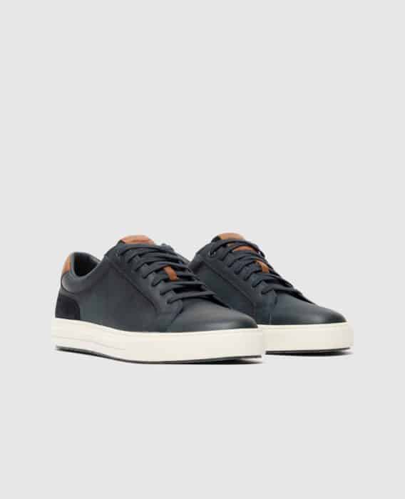 A pair of dark green low-top sneakers with white soles, black laces, and brown leather accents on the tongue and heel, displayed on a light gray background.