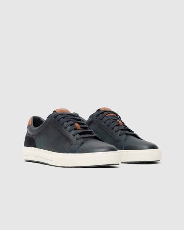 A pair of dark green low-top sneakers with white soles, black laces, and brown leather accents on the tongue and heel, displayed on a light gray background.