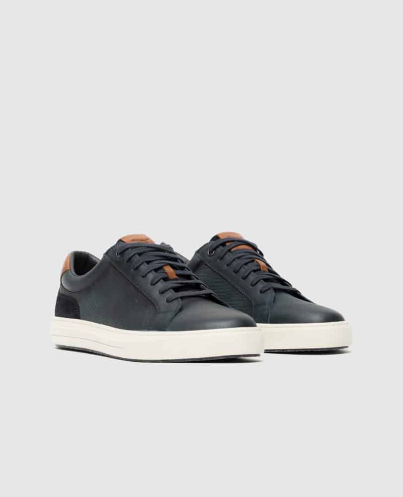A pair of dark green low-top sneakers with white soles, black laces, and brown leather accents on the tongue and heel, displayed on a light gray background.