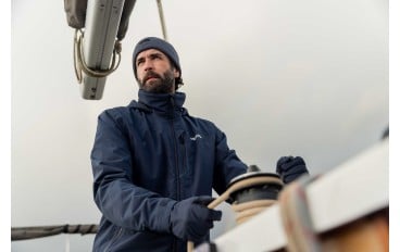 A man wearing a navy jacket, gloves, and a beanie is steering a sailboat, looking focused as he stands at the helm with one hand on the wheel and the other on a rope. The sky appears overcast.