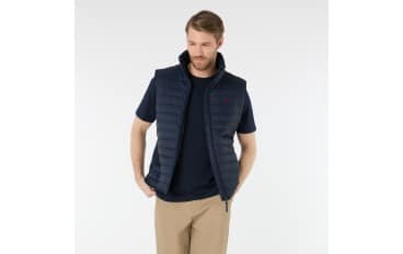 A man wearing a navy blue short-sleeve t-shirt, a dark quilted vest, and beige pants stands with one hand in his pocket, looking to the side against a plain light background.