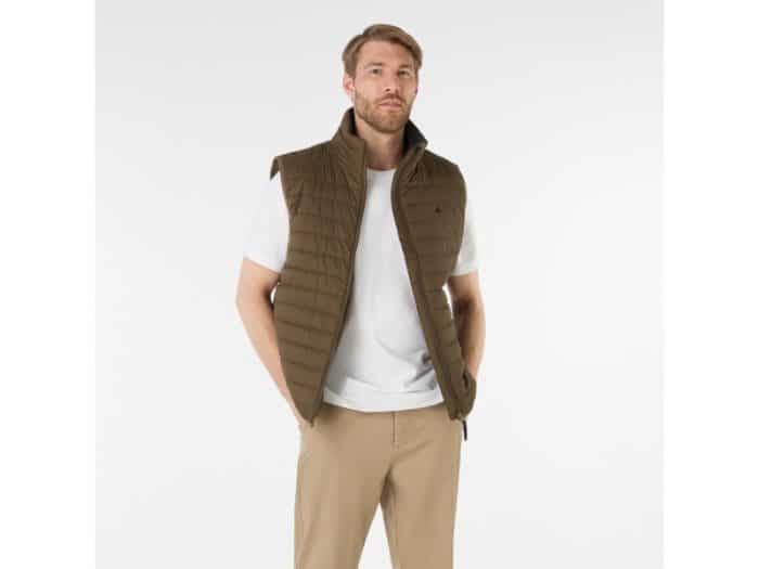 A man with light brown hair and a beard wears a white t-shirt, khaki pants, and a brown quilted vest. He stands with his hands in his pockets against a plain white background.