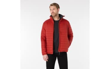 A man with short hair and a beard wears a red quilted jacket over a black shirt and black pants. He stands with one hand in his pocket against a plain white background.