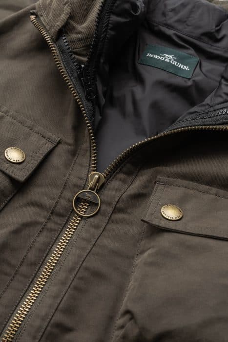 Close-up of a brown jacket with gold snap buttons and a zipper, featuring a Rodd & Gunn label on the inner collar. The fabric appears durable and the jacket has a high collar design.