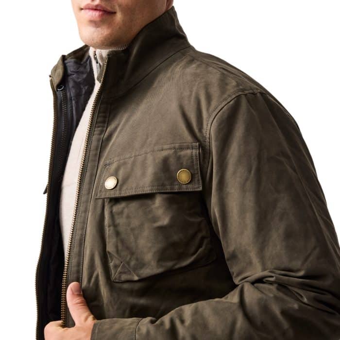 A man wearing an olive green jacket with a high collar, snap-button chest pocket, and zipper closure over a light-colored sweater. The photo is cropped, showing only the lower half of his face and torso.