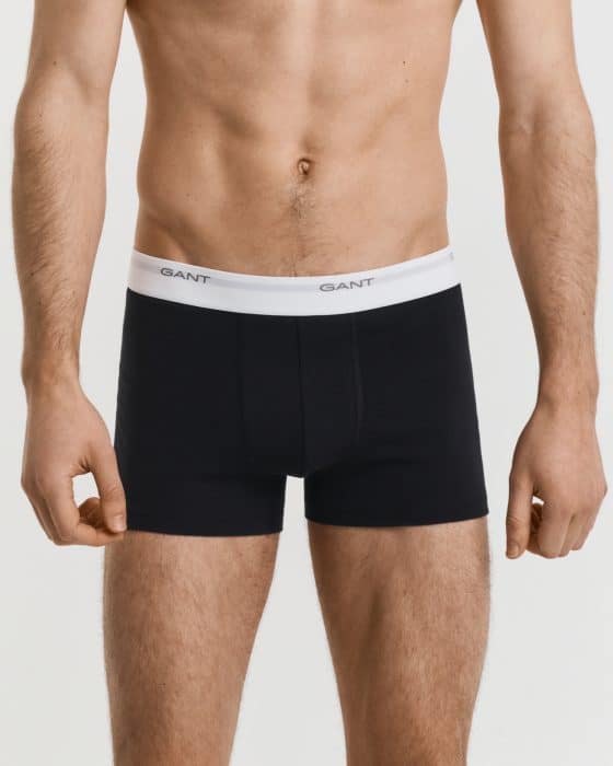 A man is standing wearing black boxer briefs with a white elastic waistband featuring the brand name GANT in black lettering. The background is plain and light-colored.