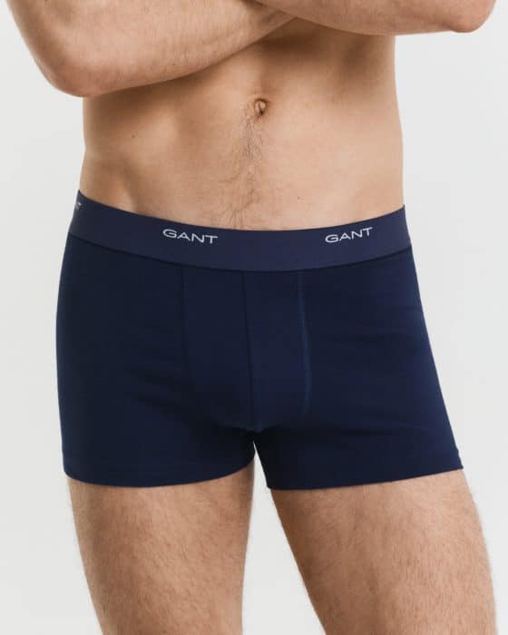 A man wearing navy blue GANT boxer briefs stands with his arms crossed. The underwear has a dark waistband with the GANT logo in white. Only his torso and upper thighs are visible on a plain white background.