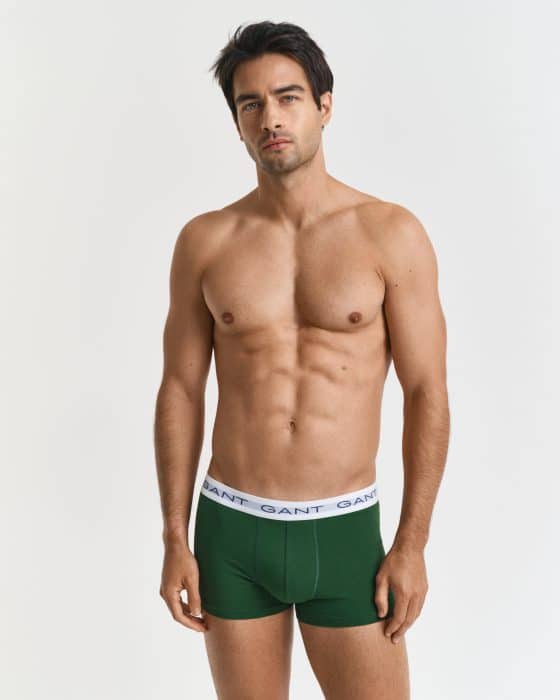 A fit, young man with short dark hair stands against a plain white background, wearing green boxer briefs with a white waistband that reads GANT. He is looking directly at the camera with a neutral expression.