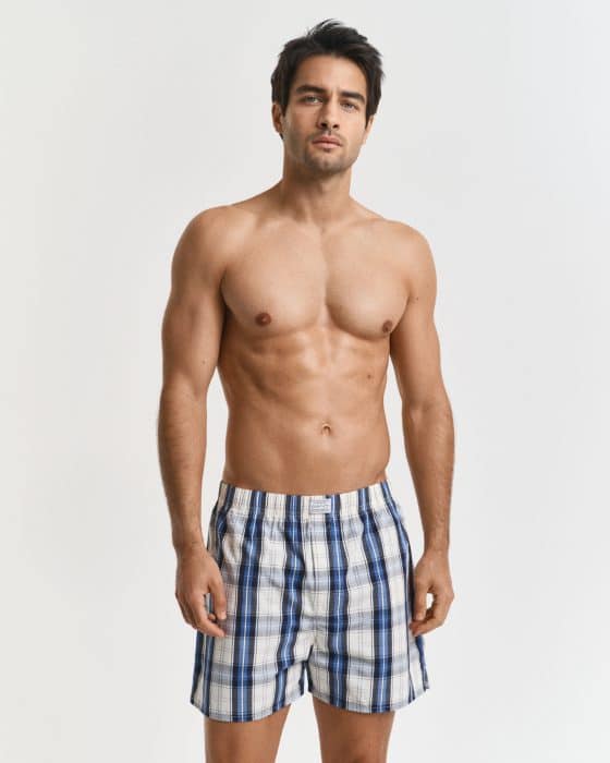 A shirtless man stands against a plain white background, wearing blue and white plaid boxer shorts.