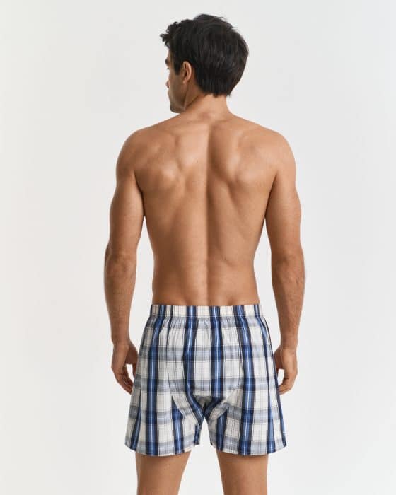 A man stands with his back facing the camera, wearing blue and white plaid boxer shorts. He is shirtless and positioned against a plain white background.