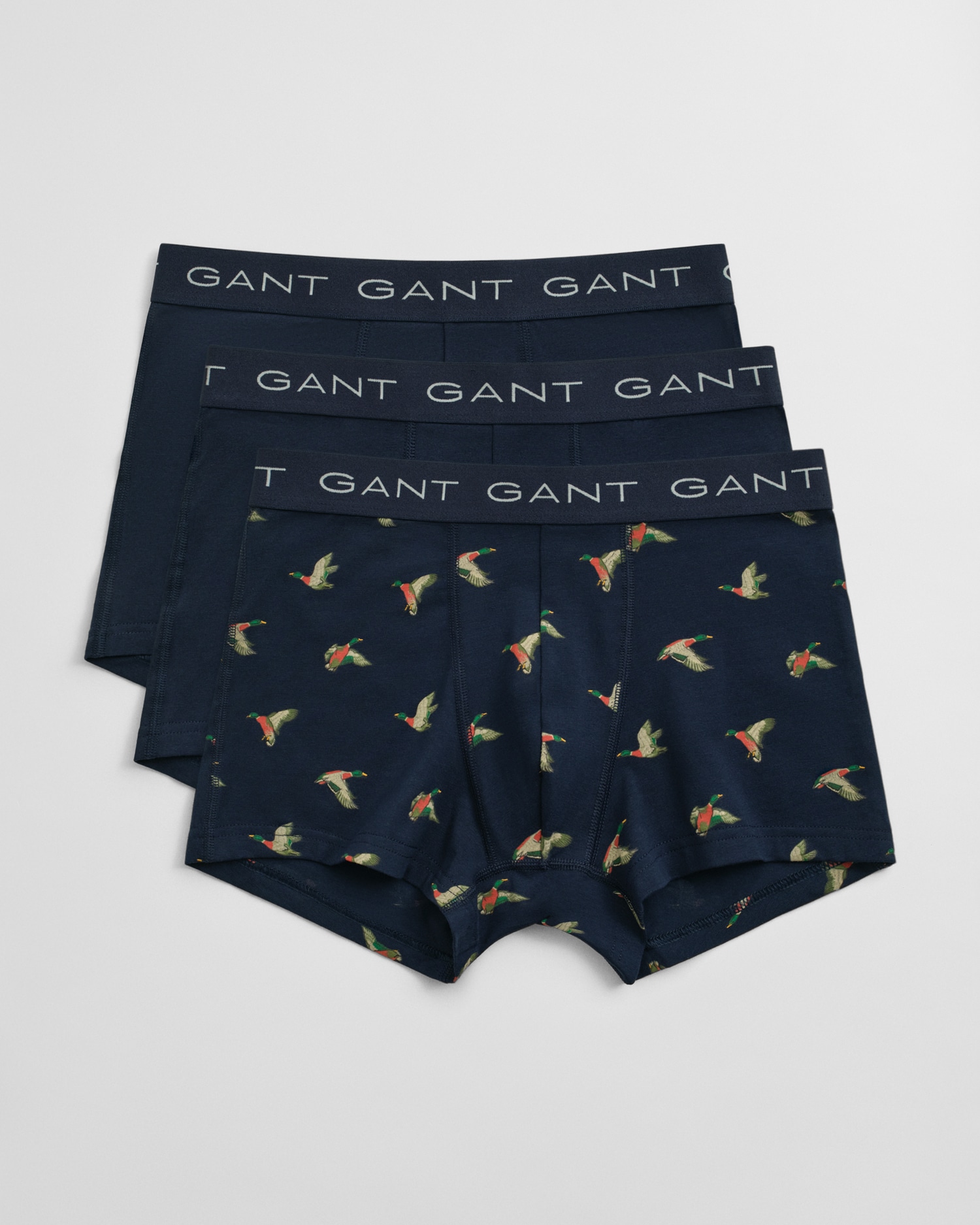 Three pairs of men’s boxer briefs are arranged in a stack. Two pairs are solid navy blue, and the top pair is navy blue with a colorful parrot print. All have GANT written on the waistband.