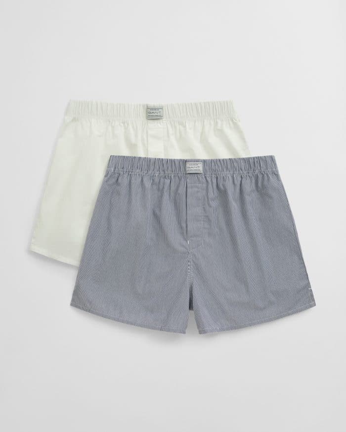 Two pairs of mens boxer shorts are displayed; one is solid white and the other is light blue with subtle stripes. Both have elastic waistbands and labels on the front.