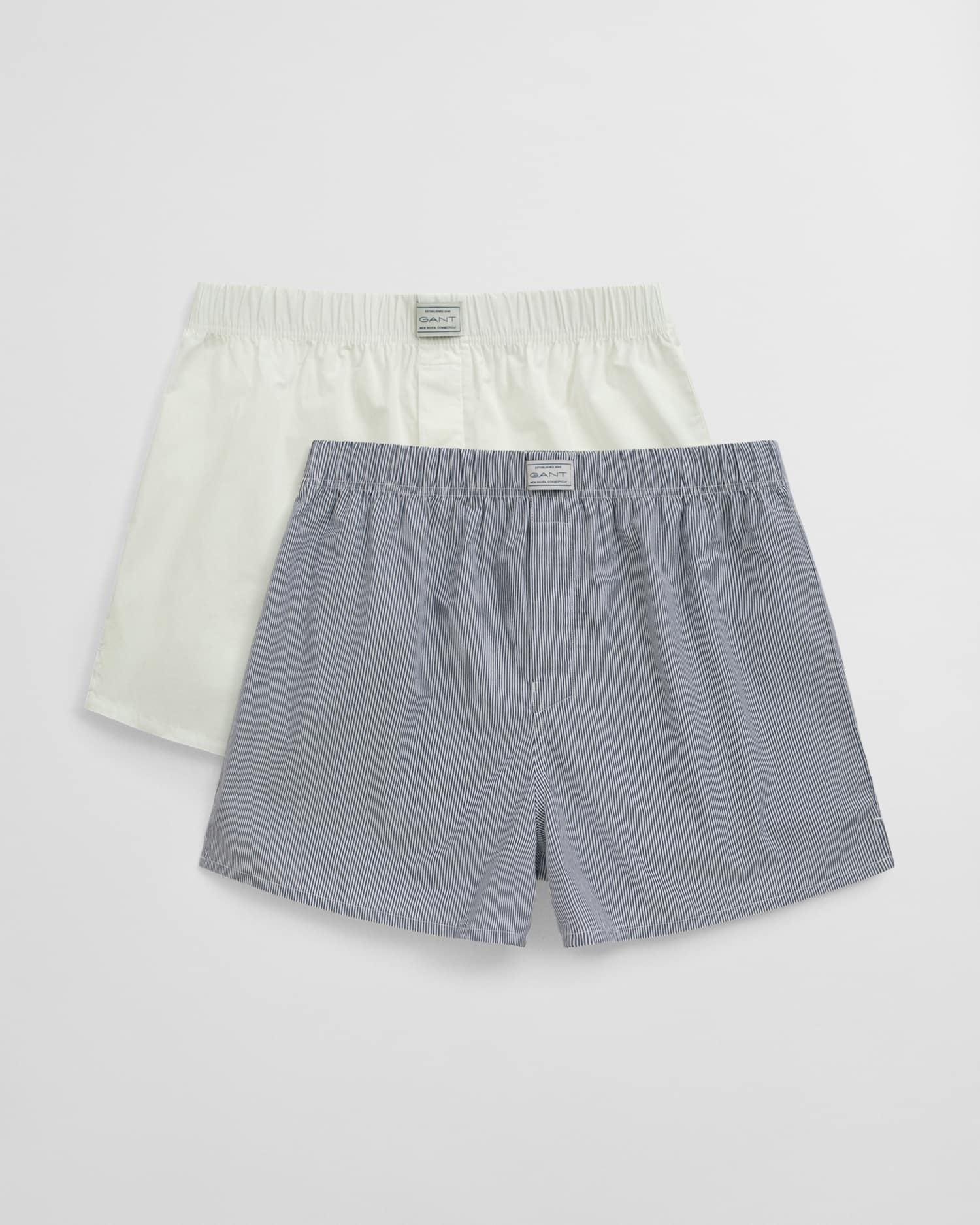 Two pairs of mens boxer shorts are displayed; one is solid white and the other is light blue with subtle stripes. Both have elastic waistbands and labels on the front.