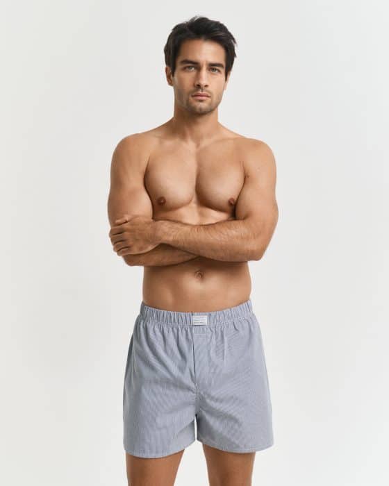 A man with dark hair stands against a plain white background, shirtless, wearing light gray boxer shorts and crossing his arms over his chest.