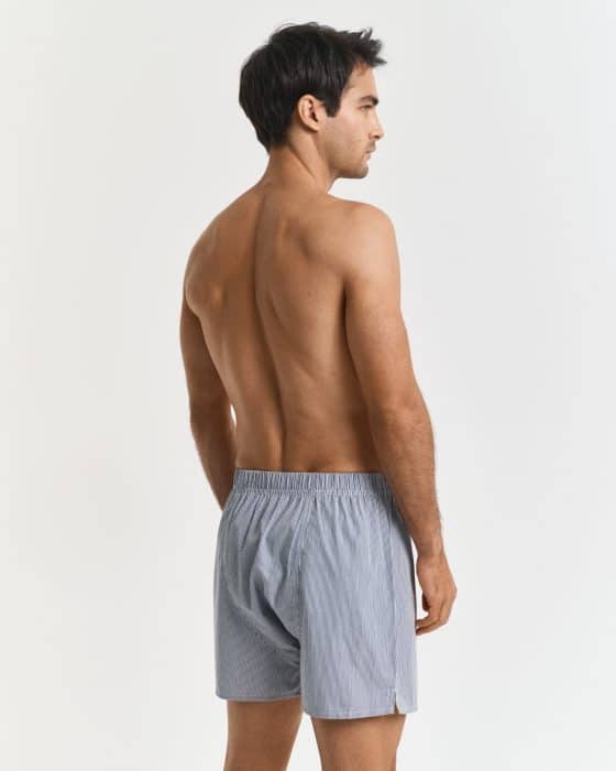 A man with short dark hair, seen from the back and side, stands shirtless wearing light blue boxer shorts against a plain white background.