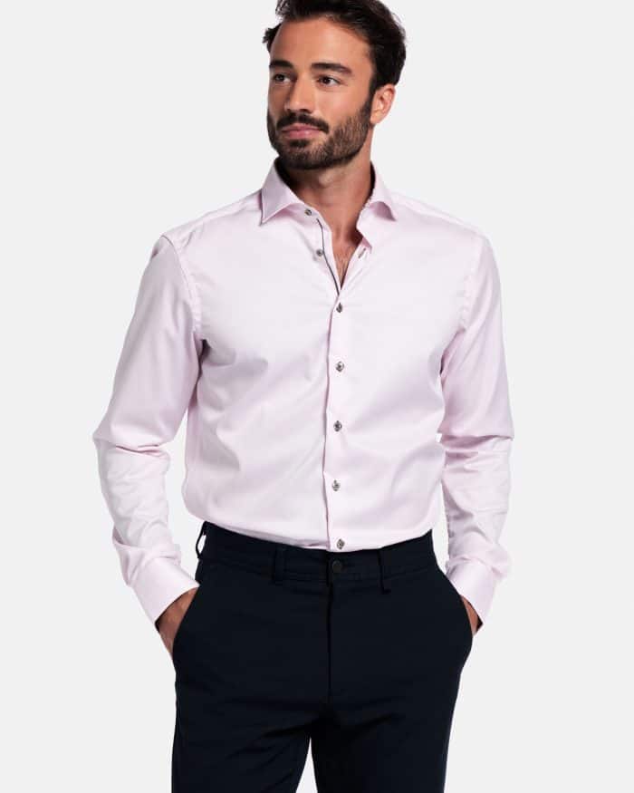 A man with short dark hair and a beard is wearing a light pink button-up dress shirt and dark trousers. He has his hands in his pockets and is looking to the side against a plain white background.
