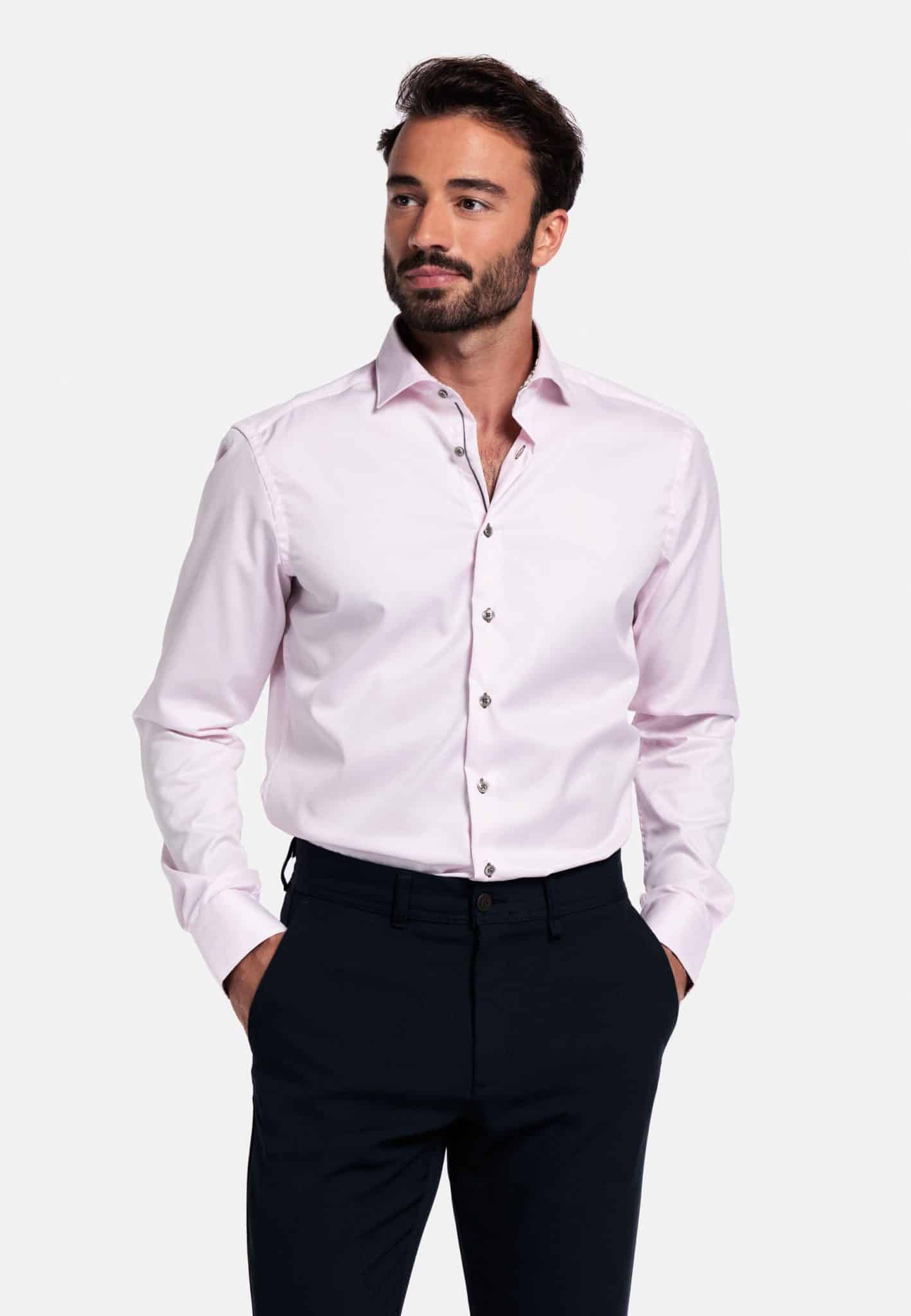 A man with short dark hair and a beard is wearing a light pink button-up dress shirt and dark trousers. He has his hands in his pockets and is looking to the side against a plain white background.
