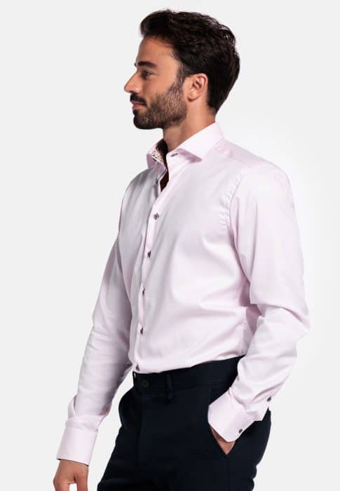 A man with dark hair and a beard is standing sideways, wearing a light pink dress shirt and dark trousers. The image is set against a plain white background.