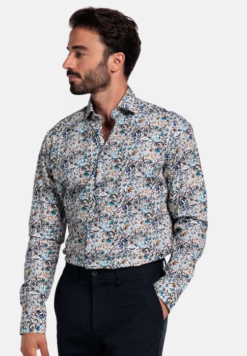 A man with dark hair and a beard wears a long-sleeve, multicolored floral print shirt tucked into dark trousers. He is standing and looking to his left against a plain light background.