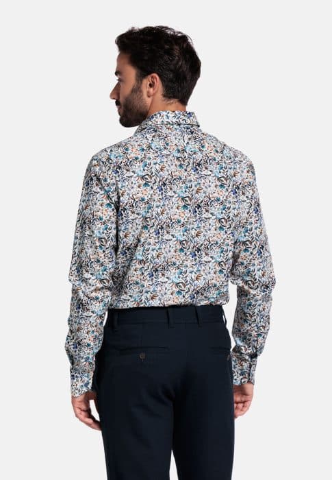 A man is standing with his back to the camera, wearing a long-sleeved, patterned floral shirt and dark dress pants against a plain white background.