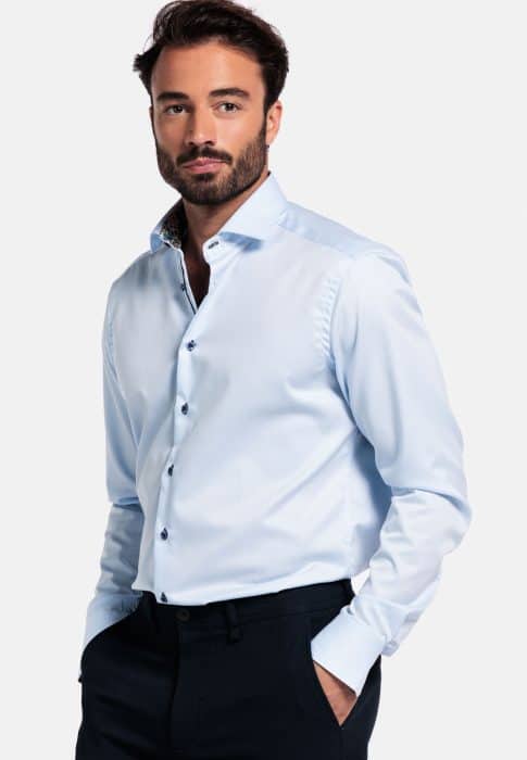 A man with dark hair and a beard wearing a light blue dress shirt and dark pants stands with his hands in his pockets, looking confidently at the camera against a plain white background.