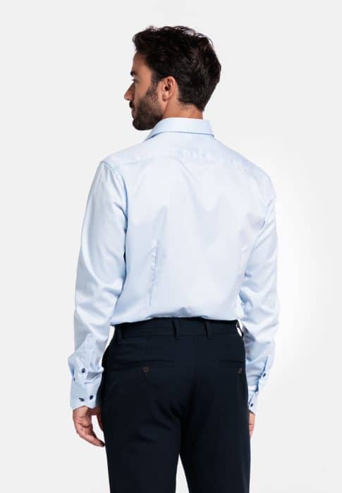 A man with dark hair is shown from the back, wearing a light blue dress shirt and dark trousers, standing against a plain white background.
