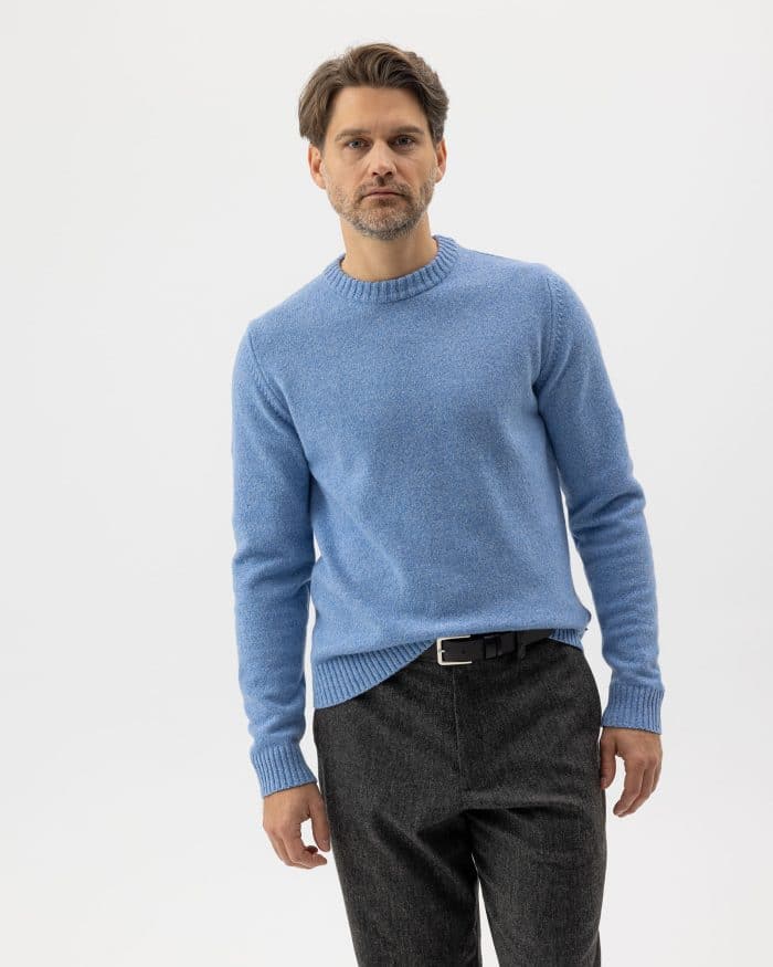 A man with light brown hair and a short beard wears a light blue sweater and dark gray pants. He stands against a plain white background, looking directly at the camera with a neutral expression.