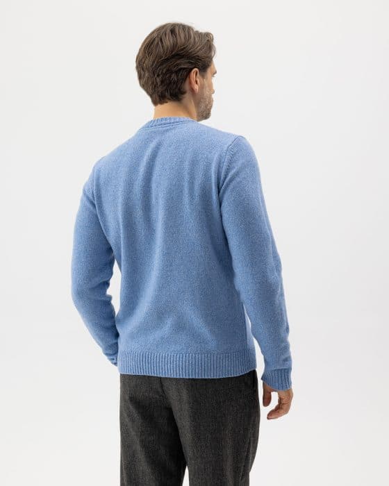 A man with brown hair, seen from the back, is wearing a light blue sweater and dark gray pants. He stands against a plain, light background.