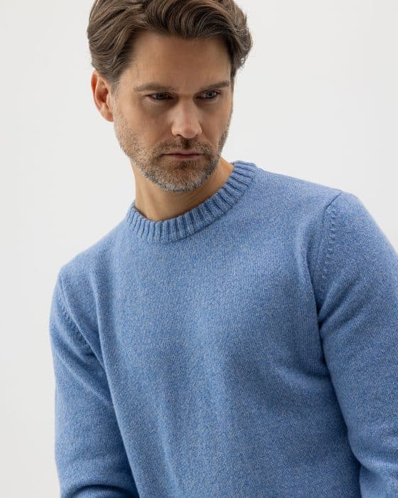 A man with brown hair and a short beard is wearing a light blue knit sweater. He is looking down with a neutral expression against a plain, light background.