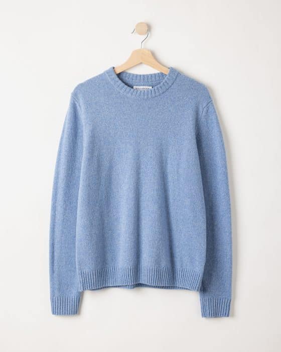 A light blue, long-sleeve crewneck sweater hangs on a wooden hanger against a plain white wall.