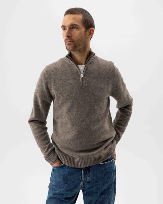 A man in a brown zip-up sweater and blue jeans stands with his hands in his pockets, looking off to the side against a plain white background.