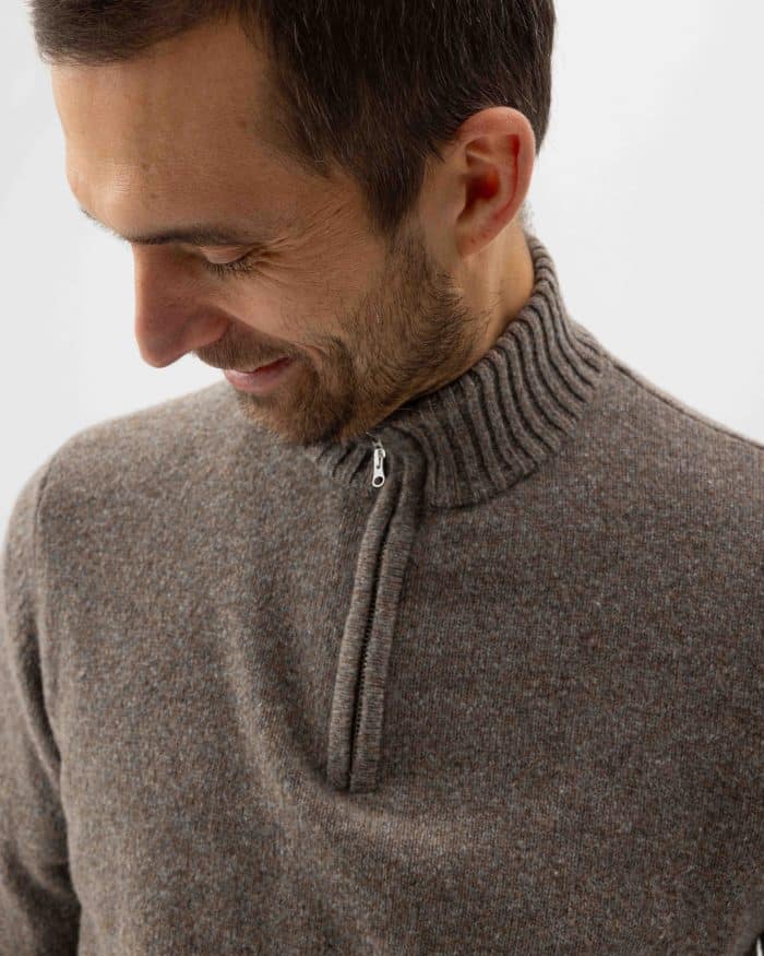 A man with short brown hair and a beard smiles while looking down. He is wearing a greyish-brown half-zip knit sweater with a ribbed collar. The background is plain and white.