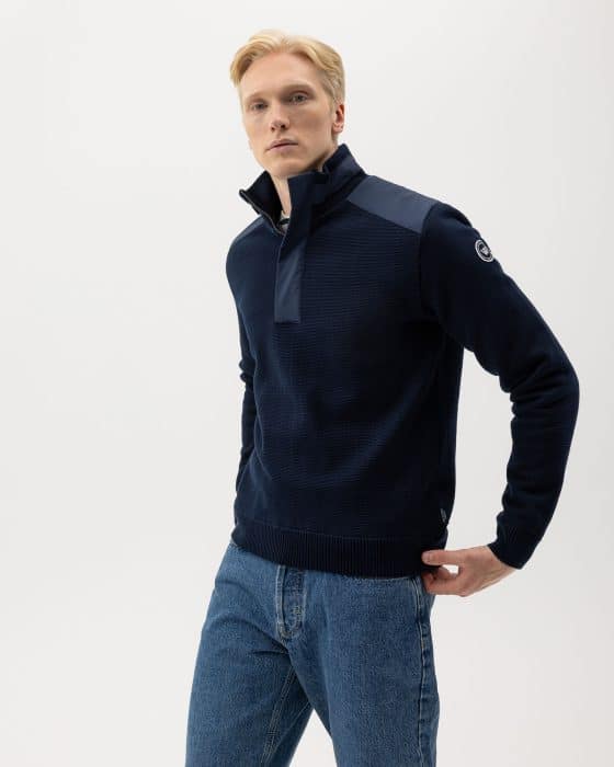 A man with short blond hair wears a navy quarter-zip sweater with dark patches on the shoulders and blue jeans, standing against a plain white background.
