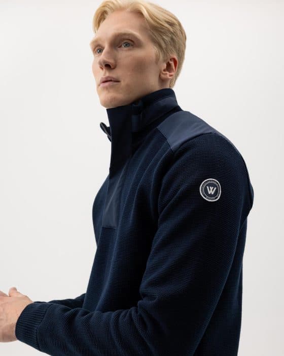 A blonde man wearing a dark blue zip-up sweater with a logo patch on the sleeve stands against a plain white background, looking slightly upward and to the side.