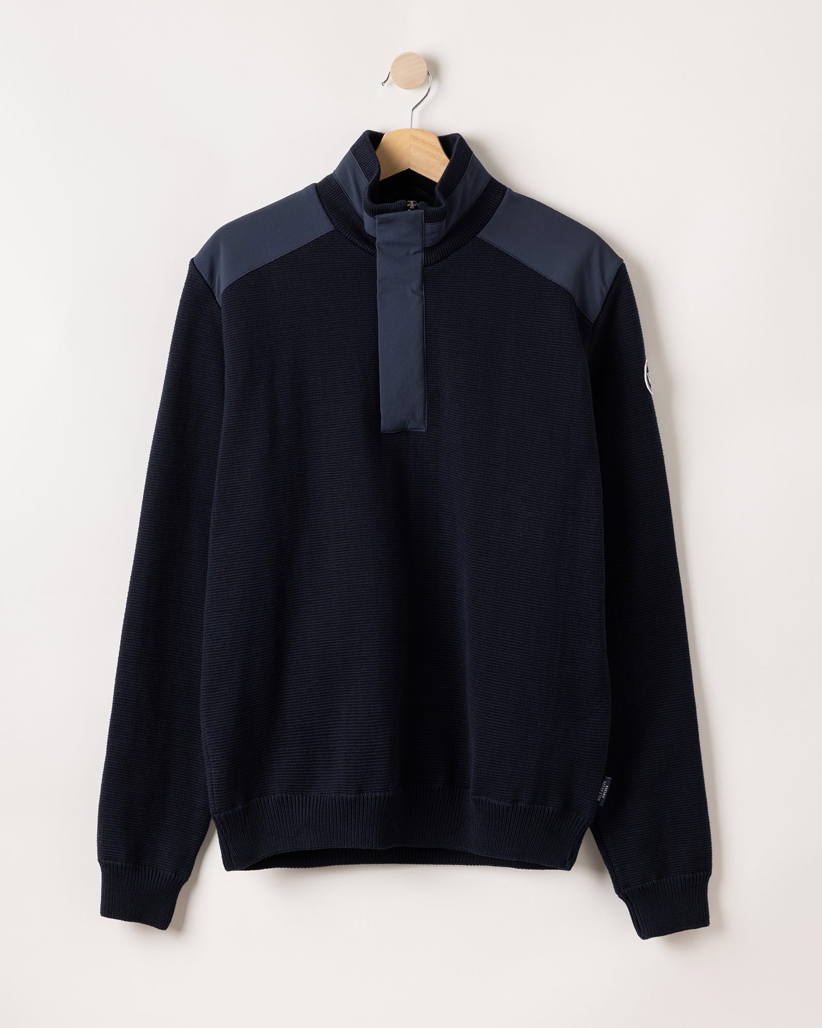 A dark navy long-sleeve pullover with a high collar, buttoned placket, and contrasting dark blue shoulder panels, displayed on a wooden hanger against a light background.
