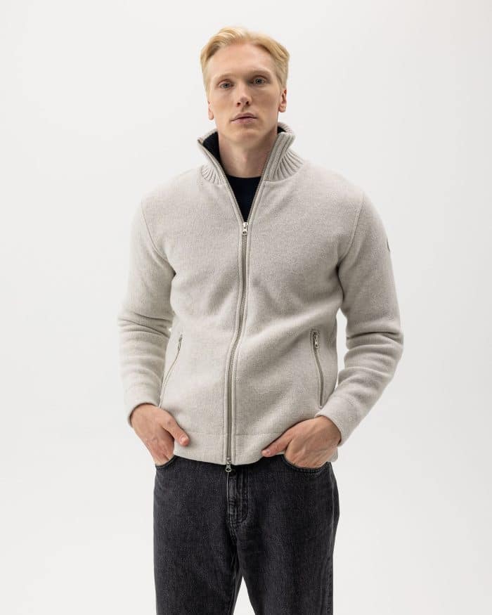 A man with light hair wearing a light gray zip-up sweater and dark jeans stands against a plain white background, looking directly at the camera with his hands in his pockets.