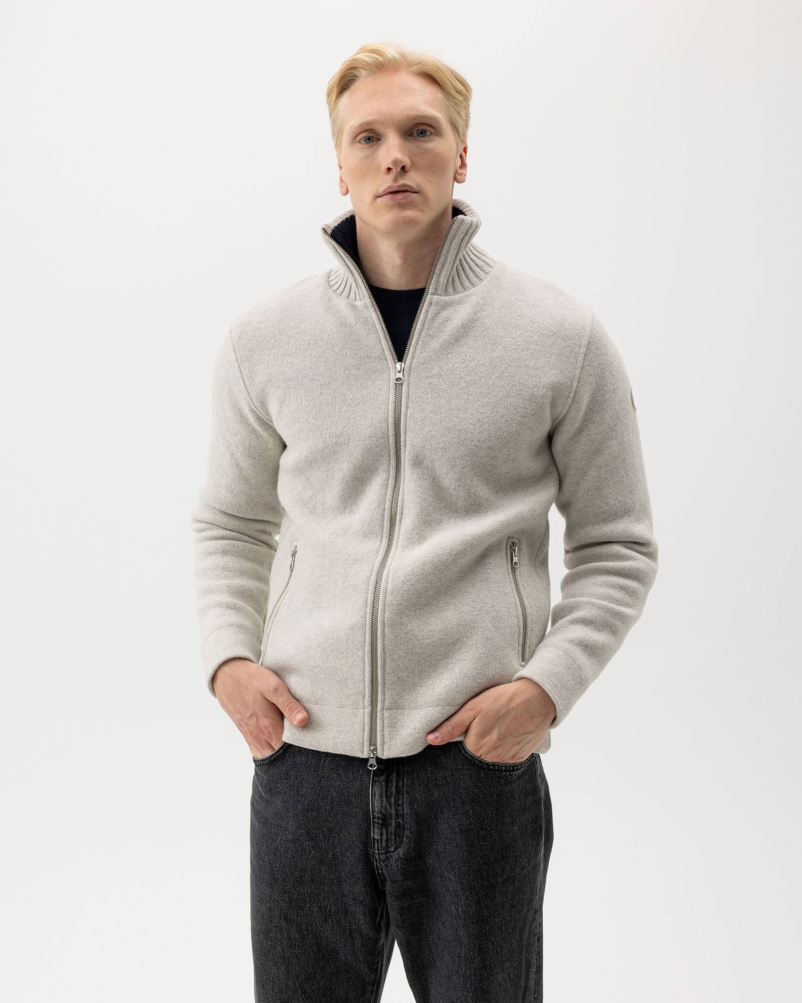 A man with light hair wearing a light gray zip-up sweater and dark jeans stands against a plain white background, looking directly at the camera with his hands in his pockets.