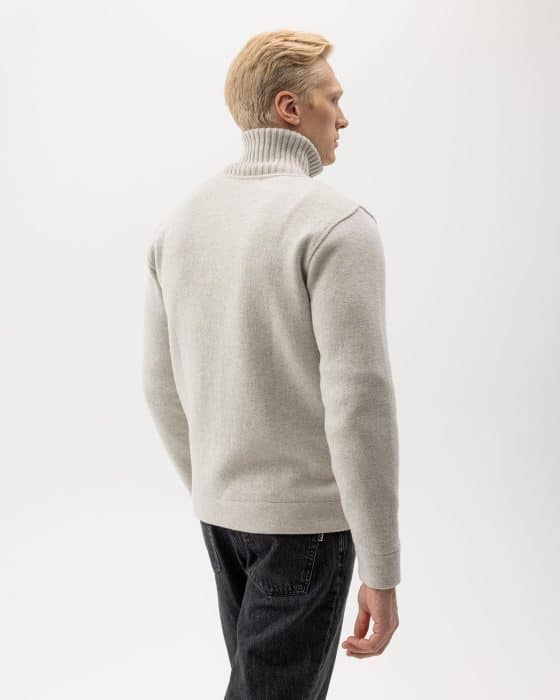 A man with short blond hair is wearing a light gray turtleneck sweater and dark jeans, facing away from the camera against a plain white background.