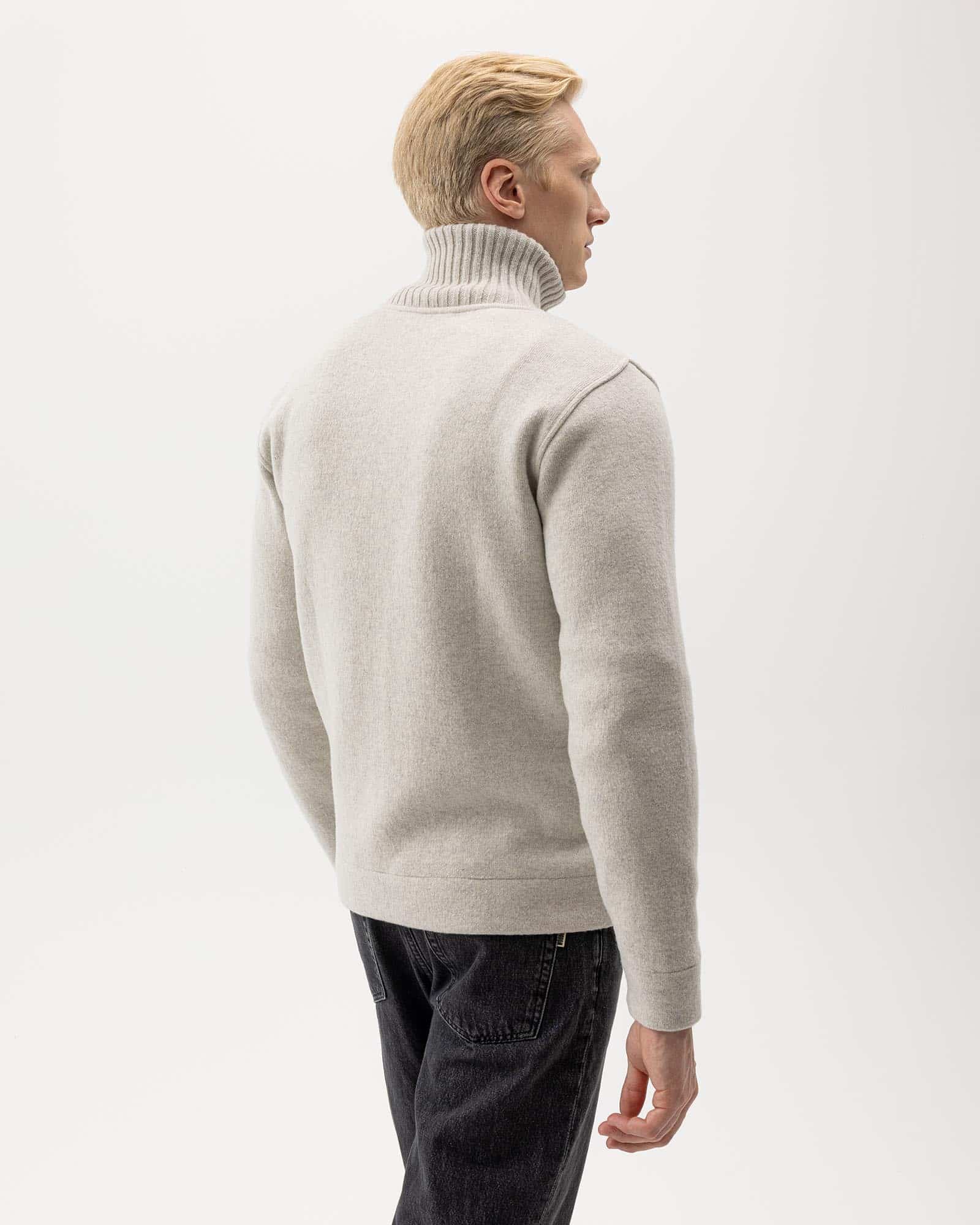 A man with short blond hair is wearing a light gray turtleneck sweater and dark jeans, facing away from the camera against a plain white background.