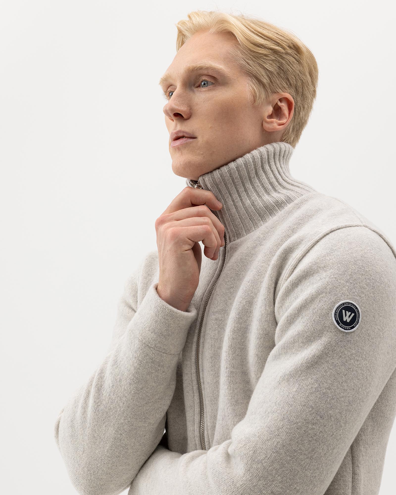 A person with short blond hair wearing a light gray zip-up sweater with a ribbed turtleneck and a circular patch on the sleeve, posing thoughtfully against a plain white background.
