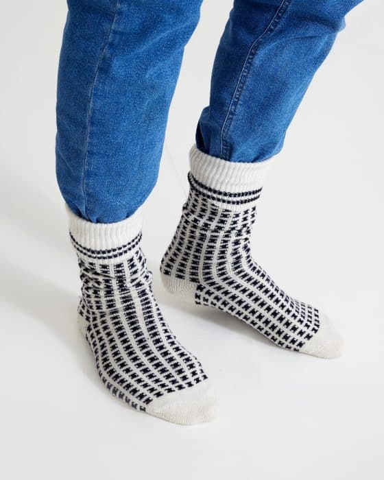 Person wearing blue jeans and white socks with a black checkered pattern, standing on a white surface. The socks are slightly slouched around the ankles.