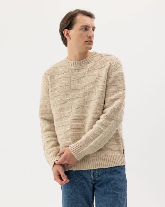 A man wearing a beige textured knit sweater and blue jeans stands against a plain white background, looking to his left with his hands clasped in front of him.
