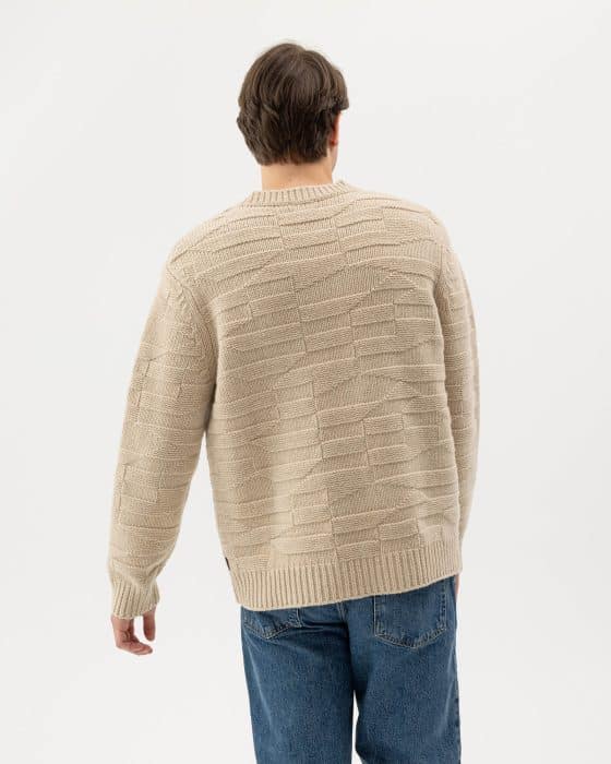 A person with brown hair is shown from the back, wearing a cream-colored textured sweater and blue jeans, standing against a plain white background.