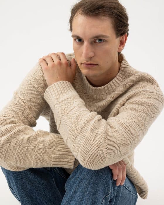 A person with long brown hair, wearing a textured beige sweater and blue jeans, sits with one knee raised, resting an arm on their knee, looking directly at the camera with a neutral expression.