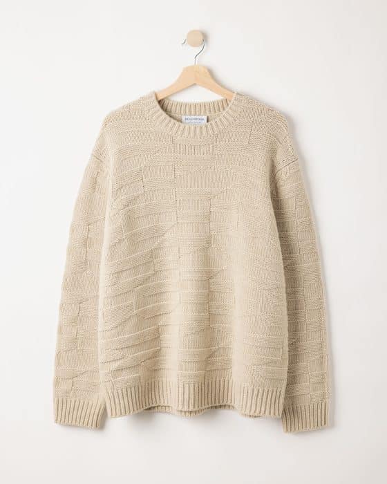 A beige knitted sweater with a textured pattern hangs on a wooden hanger against a plain white wall.