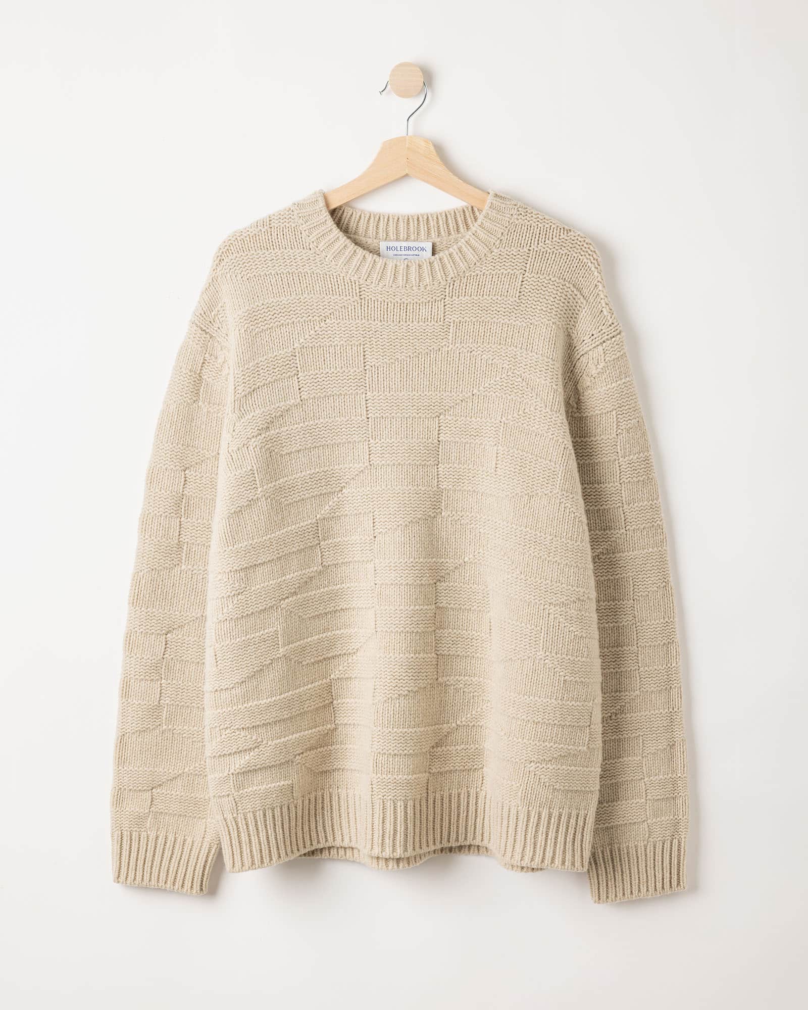 A beige knitted sweater with a textured pattern hangs on a wooden hanger against a plain white wall.