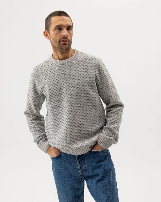 A man wearing a light gray patterned sweater and blue jeans stands with his hands in his pockets against a plain white background.