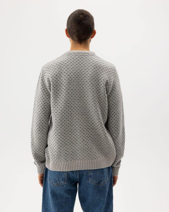 A person with short hair, seen from behind, wears a light grey textured sweater and blue jeans, standing against a plain white background.